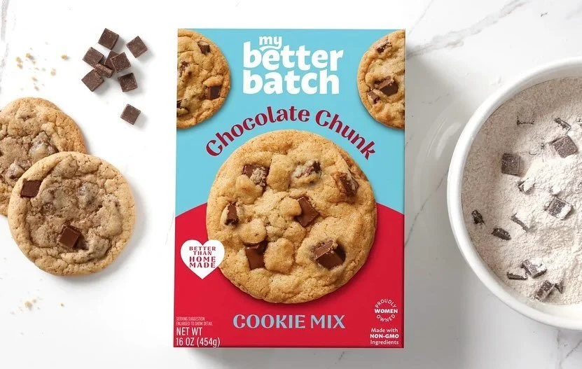 Box of Better Batch Chocolate Chunk Cookie Mix with finished cookies, chocolate chips, and cookie dough on a white surface.