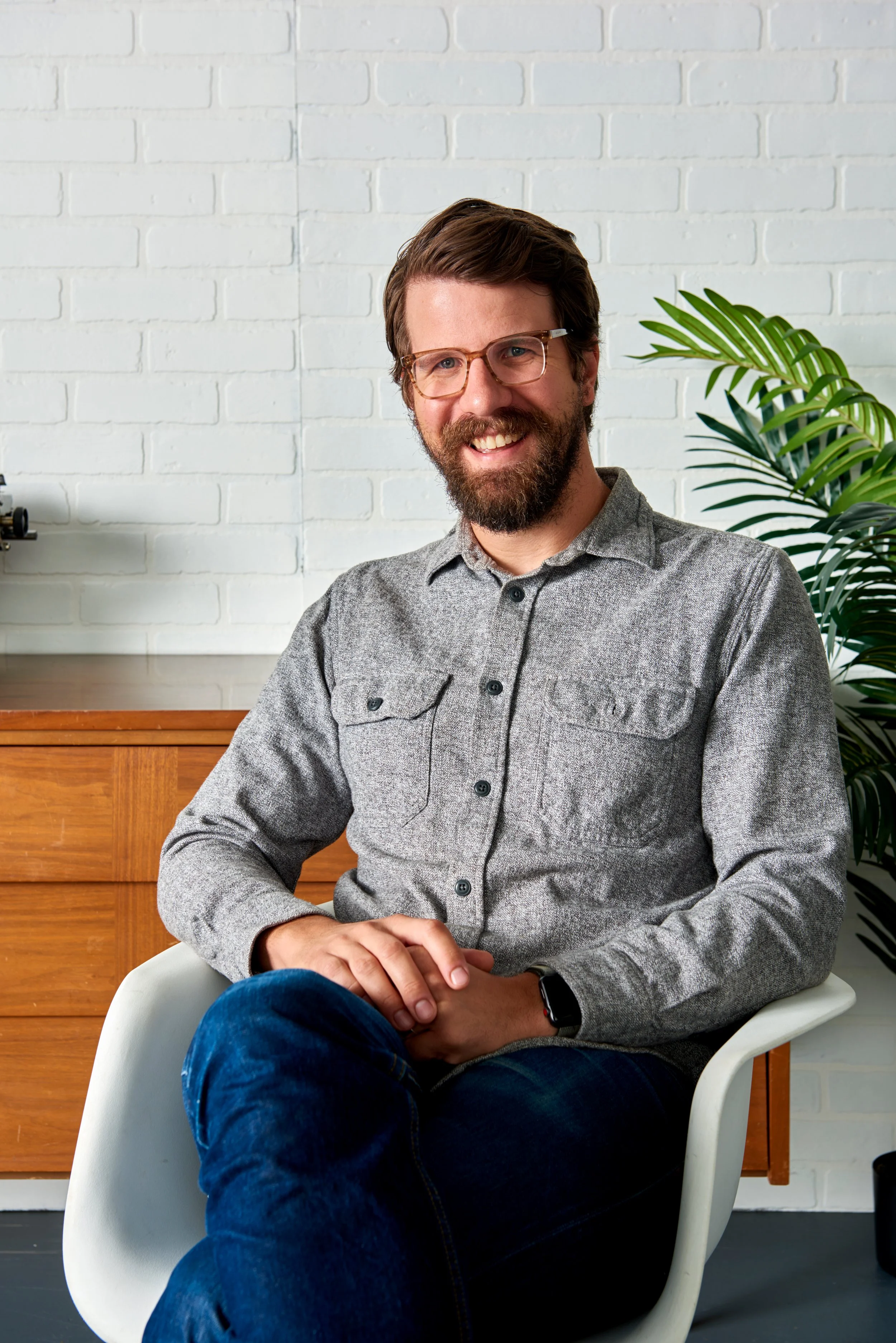 A smiling man with glasses and a beard sitting on a white chair in a modern room with a white brick wall and a green plant behind him. He is wearing a gray button-up shirt and dark blue jeans.