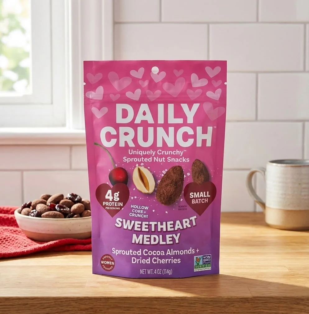 Pink bag of Daily Crunch Sweetheart Medley sprouted cocoa almonds and dried cherries on a wooden table next to a bowl of chocolate snacks and a mug.