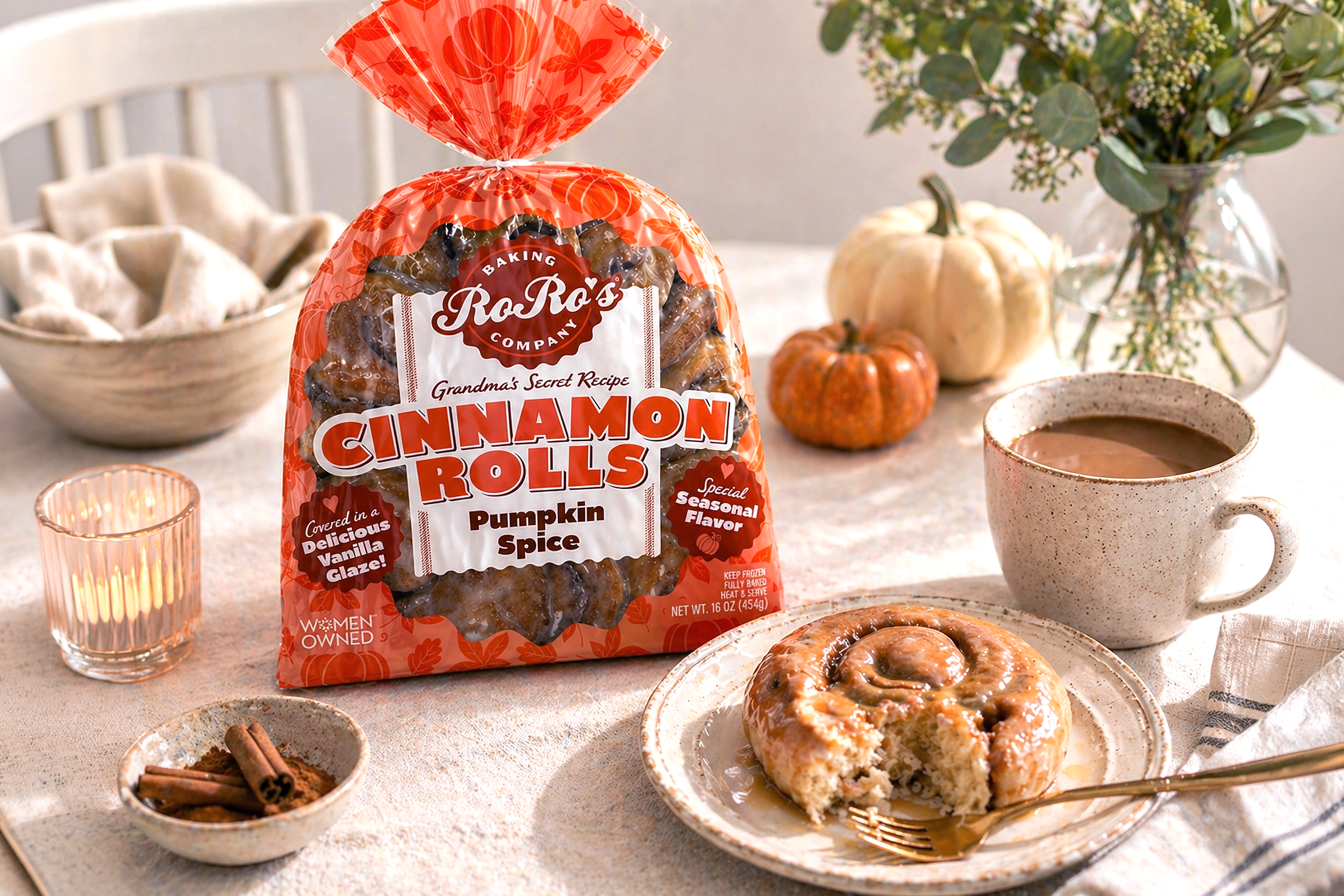 A table decorated for fall with a bag of Ro Ro's cinnamon roll pumpkin spice icing, a pumpkin, a mug of hot chocolate, a bowl of cinnamon sticks, a bowl of cookies, and a plate with a sliced cinnamon roll.