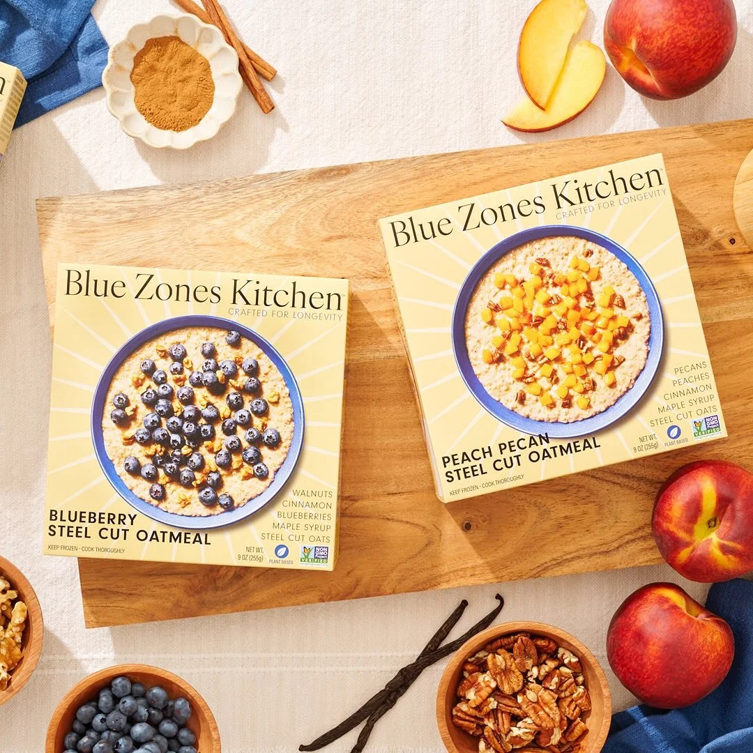 Two boxes of Blue Zones Kitchen steel cut oatmeal on a wooden board, one blueberry flavor and one peach pecan flavor, surrounded by apples, blueberries, walnuts, vanilla beans, and cinnamon sticks.