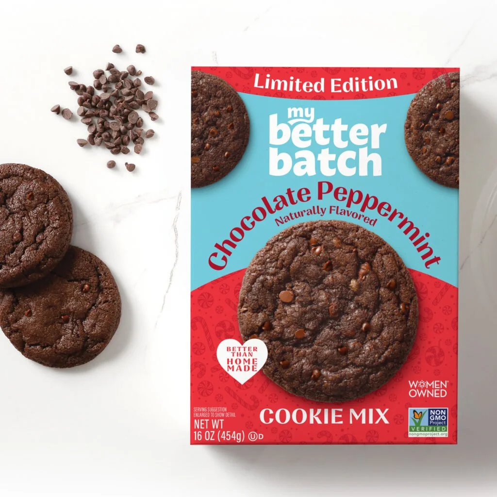 A box of Better Batch chocolate peppermint cookies flavored with chocolate chips, placed on a white surface along with three cookies and some chocolate chips.