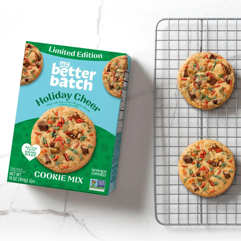 A box of Better Batch Holiday Cheer cookie mix with two cookies cooling on a wire rack, showing colorful sprinkles and chocolate chunks.