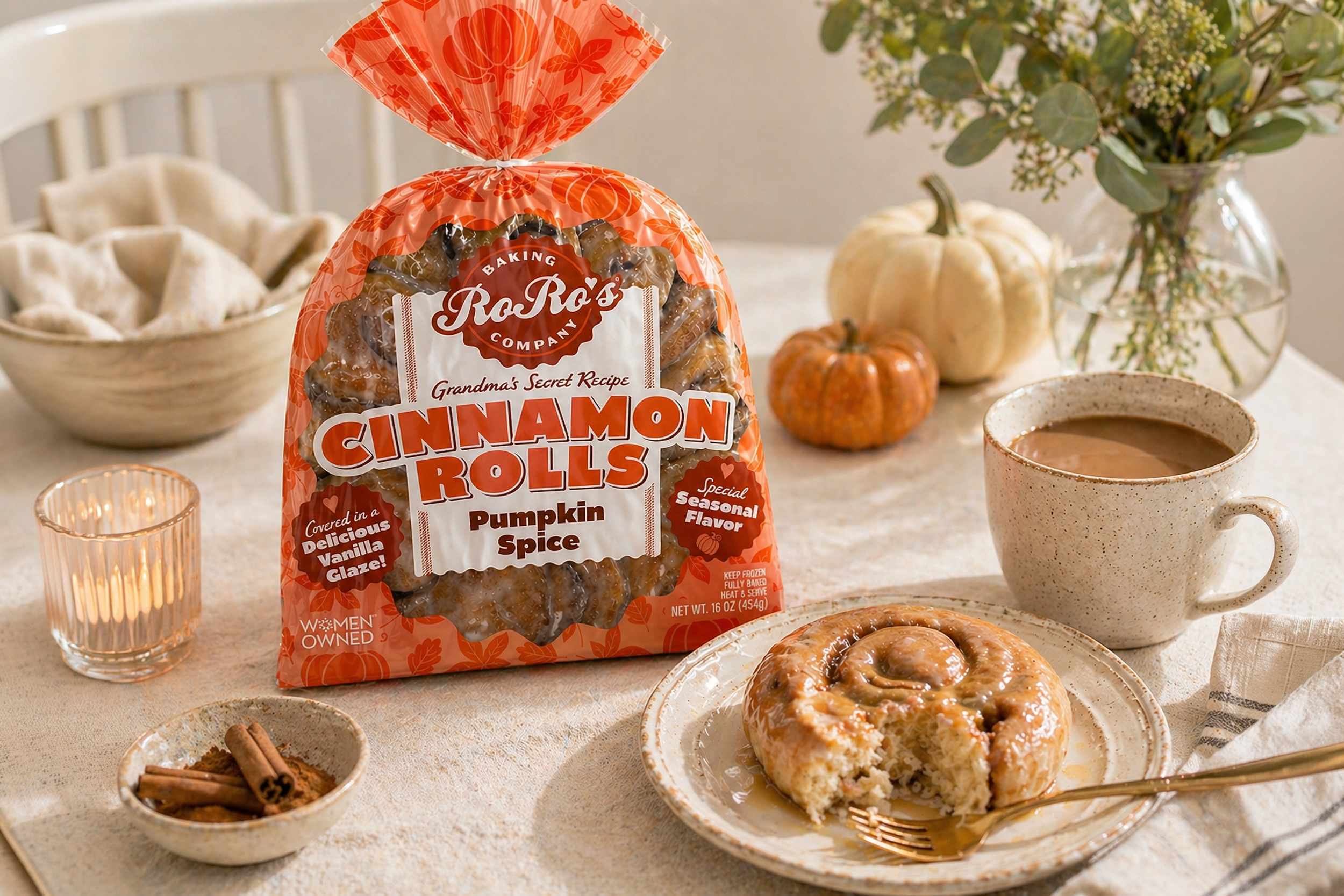Shelf with baked cinnamon rolls, a cup of coffee, a bowl of powdered sugar-coated pastries, a bag of pumpkin spice cinnamon rolls, a small bowl of cinnamon sticks, and a pumpkin and floral arrangement in the background.