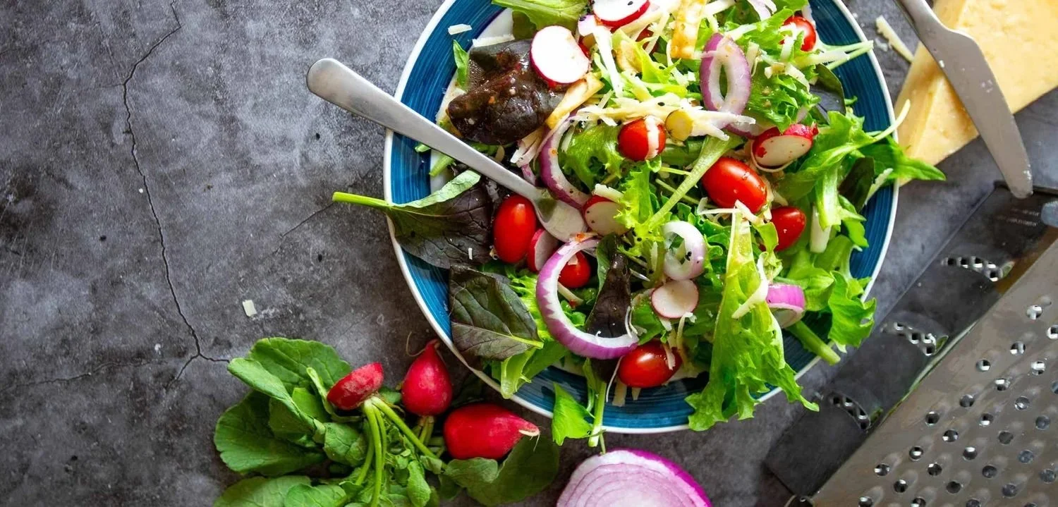 A colorful mixed salad in a blue bowl with cherry tomatoes, radishes, red onions, leafy greens, and shredded cheese, with some radishes and onion slices on the gray stone countertop.