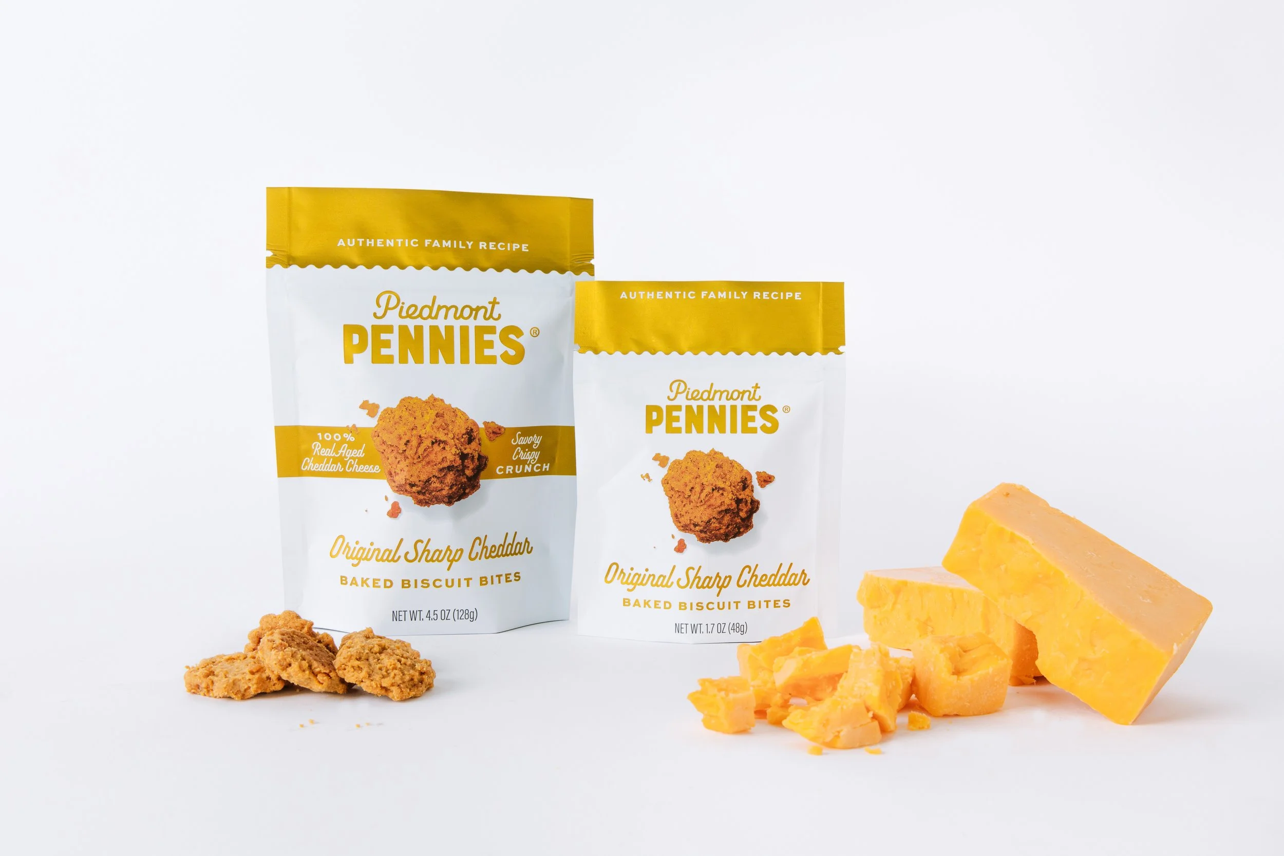 Two bags of Piedmont Pennies baked biscuit bites with cheddar cheese, along with some blocks and crumbles of cheddar cheese, set against a white background.