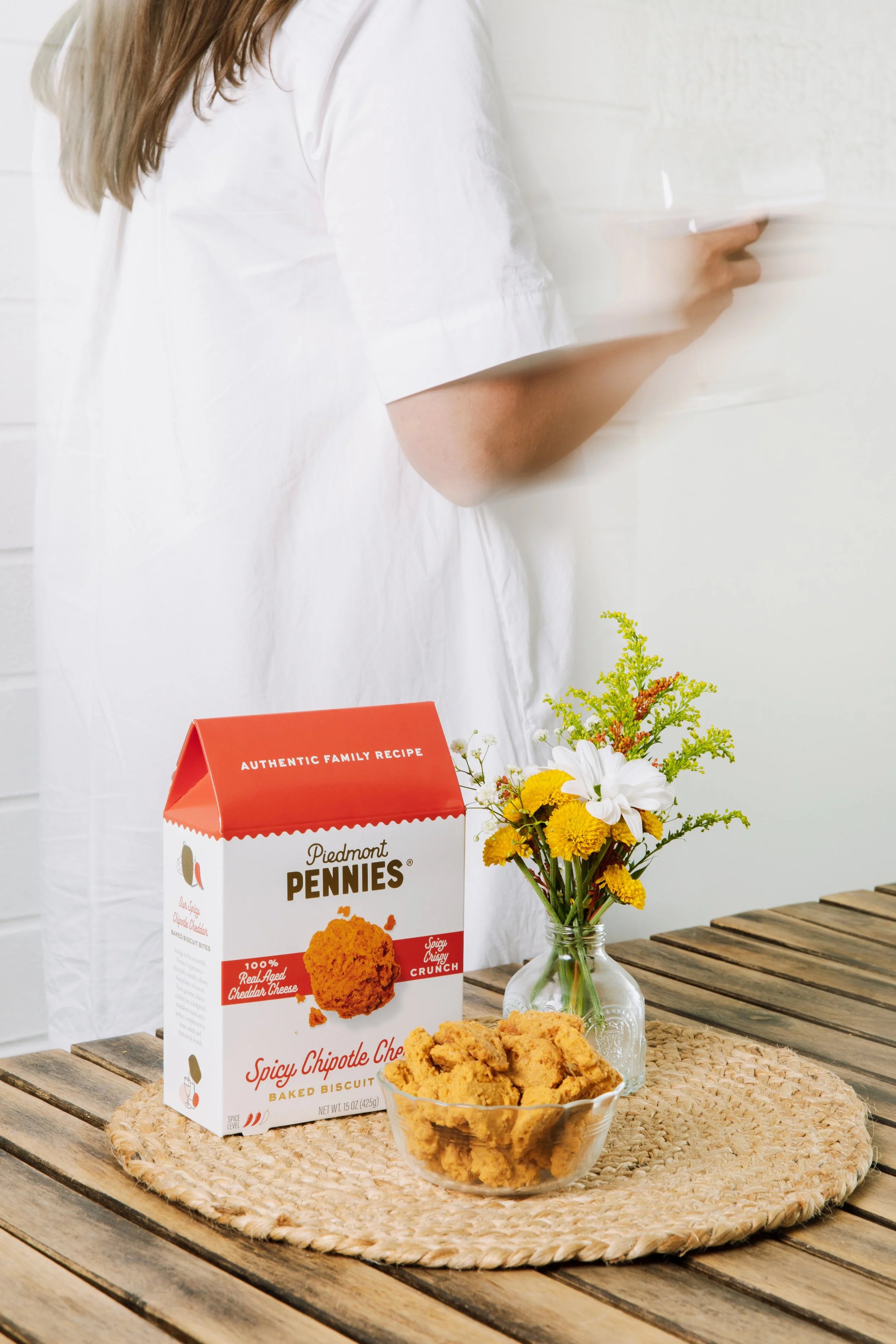 A box of Piedmont Pennies spicy chipotle chip baked biscuit bites on a woven mat on a wooden table, next to a glass jar with yellow and white flowers, with a woman in white standing behind it, holding a white container.