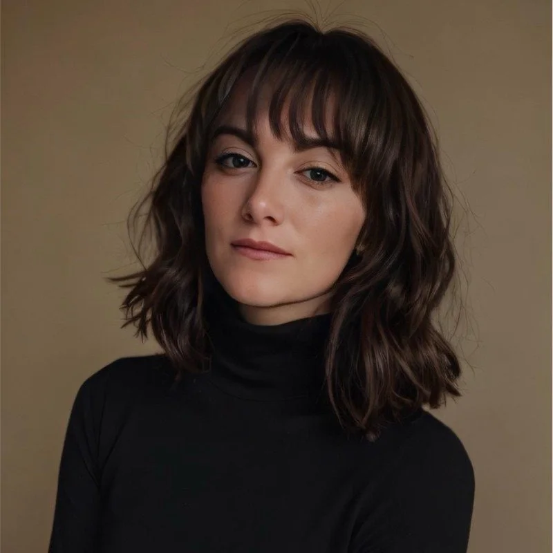 A young woman with shoulder-length wavy brown hair with bangs, wearing a black turtleneck, posing against a plain beige background.