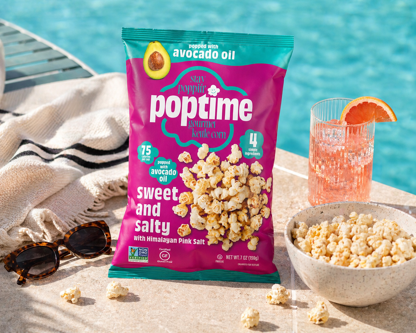 Bag of popcorn with sweet and salty Himalayan pink salt flavor, a bowl of popcorn, a glass of pink grapefruit beverage with a grapefruit slice, sunglasses, and a white towel by a poolside.