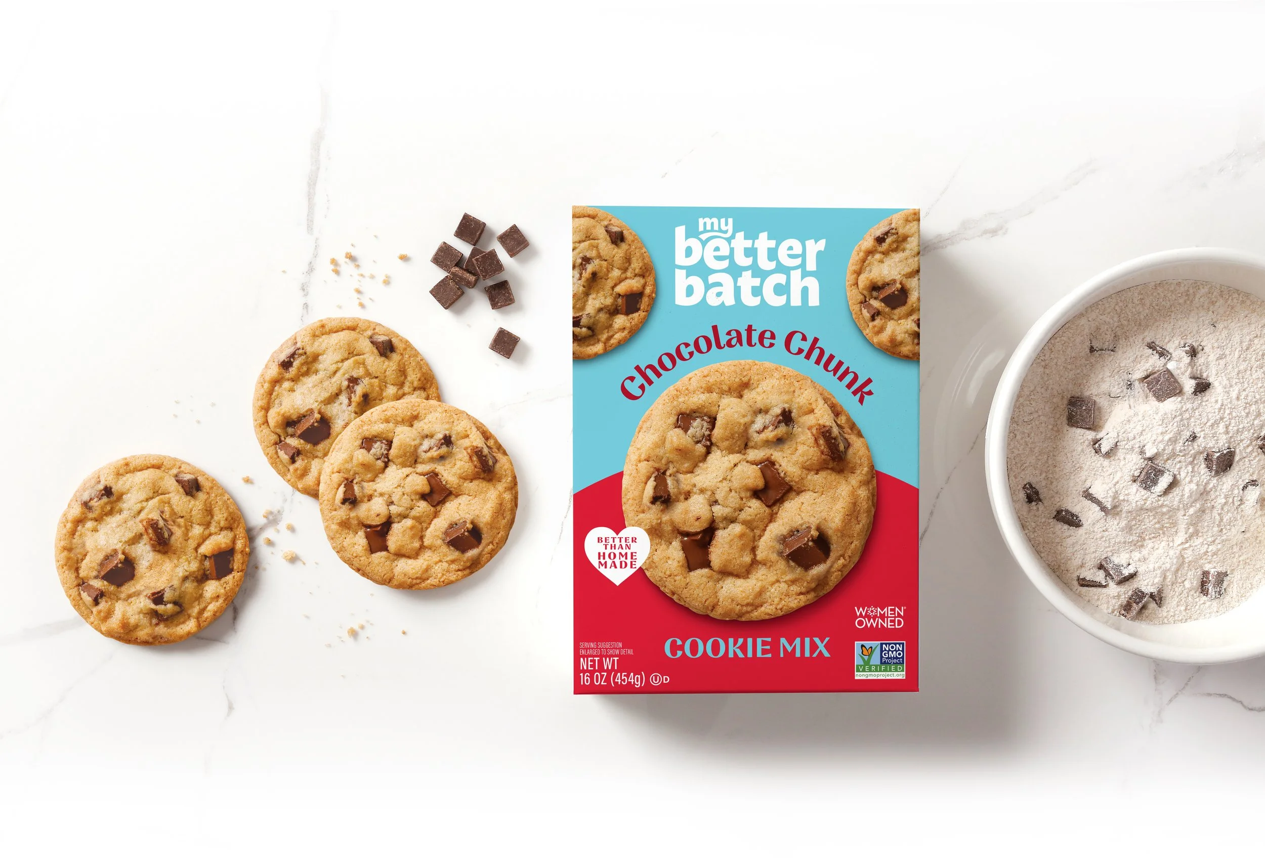Chocolate chip cookies with a box of 'my better batch' cookie mix and a cup of powdered cookie mix with chocolate chips inside, all placed on a white marble surface.