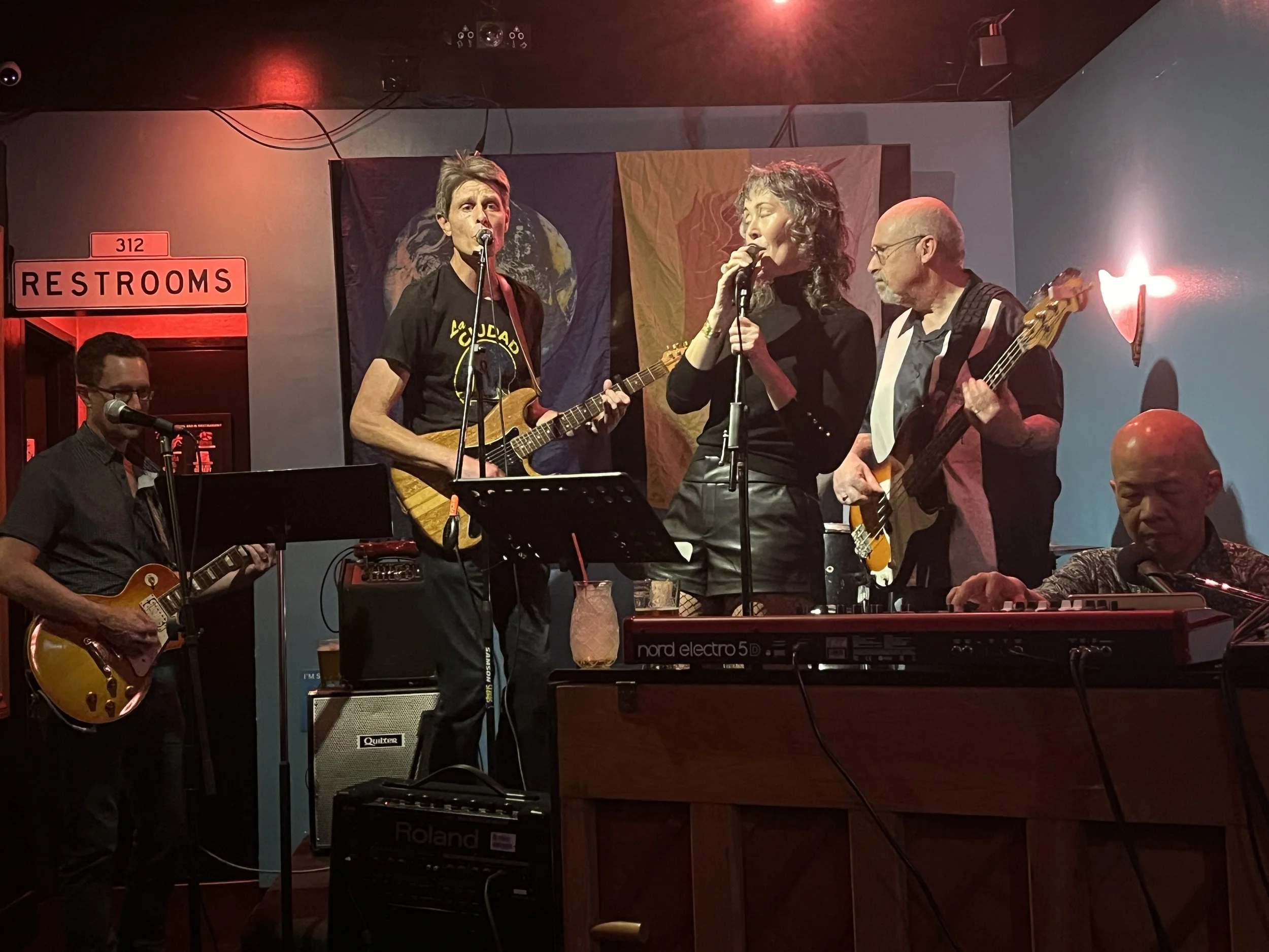 Musicians performing on a stage with red lighting at the Question Mark Bar. Three of them are playing guitars and vocals, one is playing keyboard, and three others are standing or sitting, with a 'Restrooms' sign visible to the left.