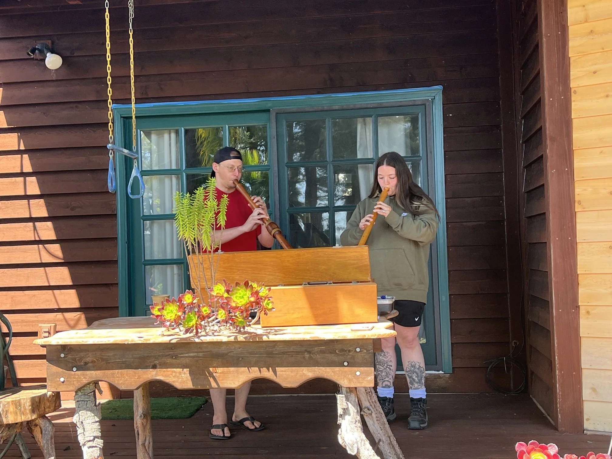Two people, Cadence for Change members, playing wooden flutes on a porch or balcony with a table holding a plant arrangement in the foreground.