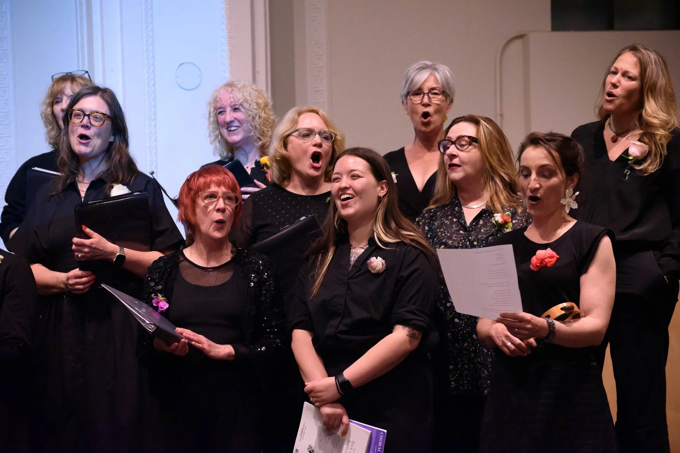 Cadence for Change members singing in choir on stage, holding sheet music, dressed in black, some with flowers and glasses, in a formal setting.
