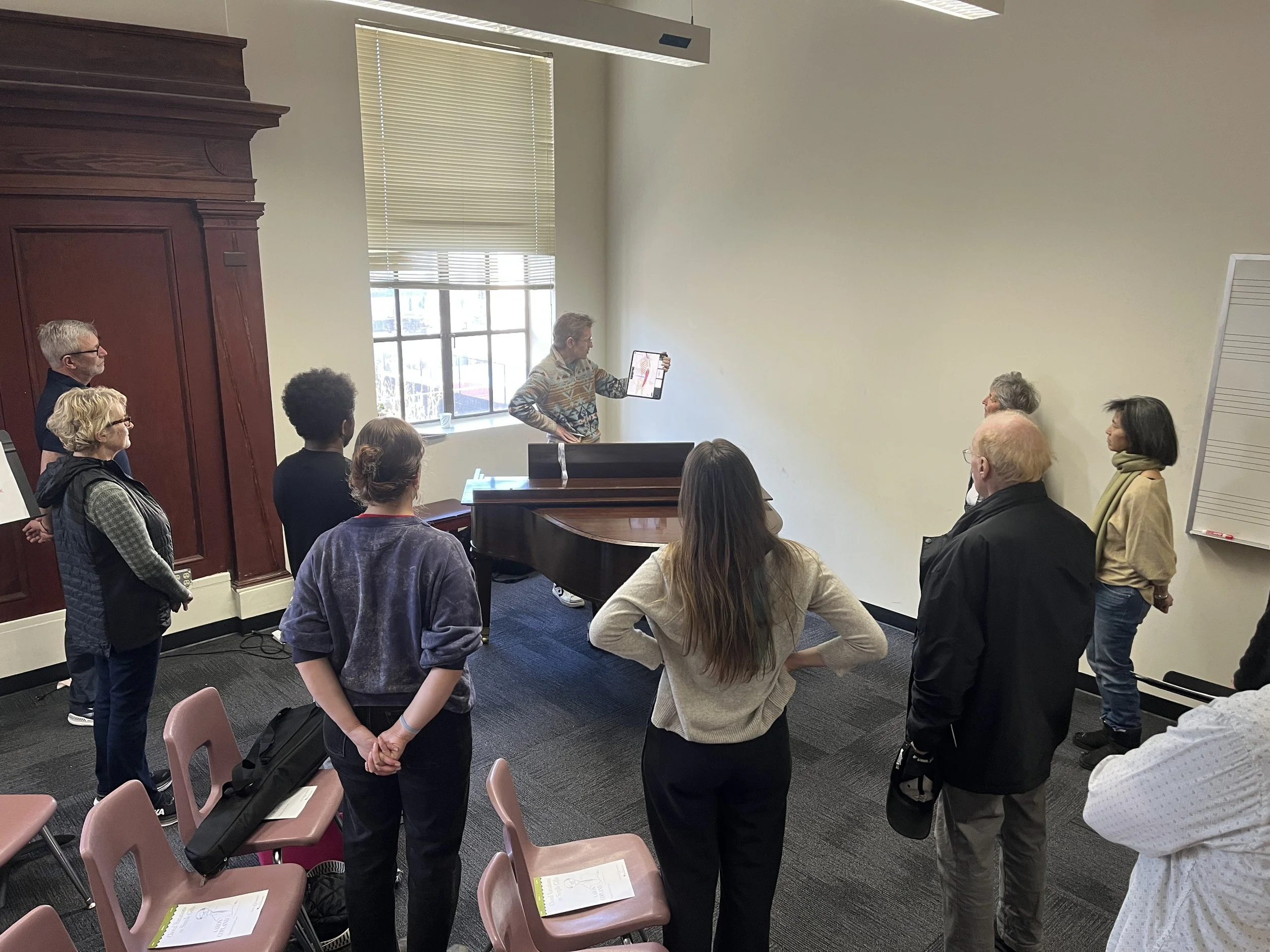Cadence for Change members gathered in a room for Voice Class, listening to a man showing a diagram on a tablet. The room has a window with blinds, a whiteboard on the wall, and chairs with papers on them.