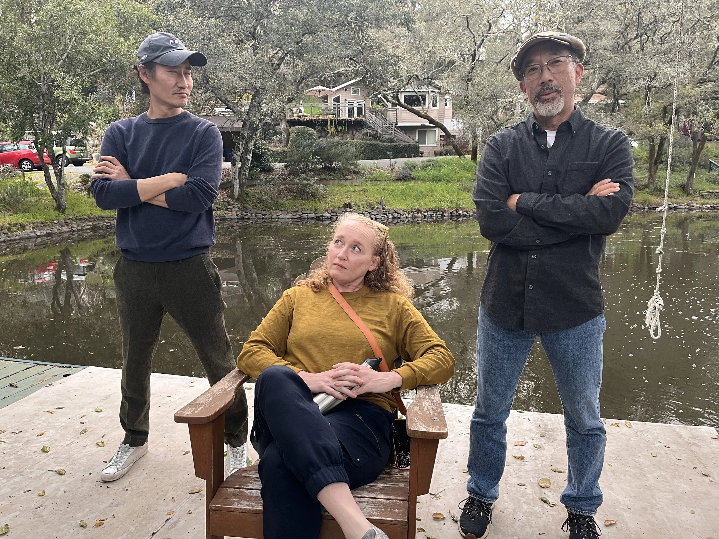 Cadence for Change members outdoors by a pond with trees and houses in the background. Two men are standing with arms crossed, and a woman is sitting on a bench looking up at one of the men. Fall leaves are on the ground.
