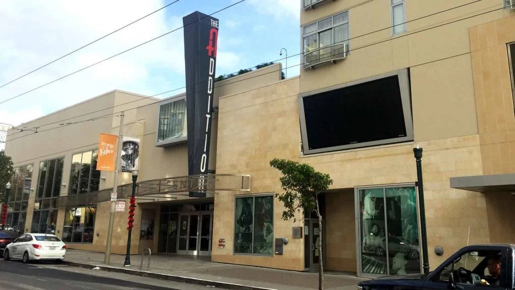 The Additions building with a vertical sign reading "THE ADDITION" in red and white letters, a large black electronic billboard, and a glass storefront, located on a city street with cars parked along the curb.