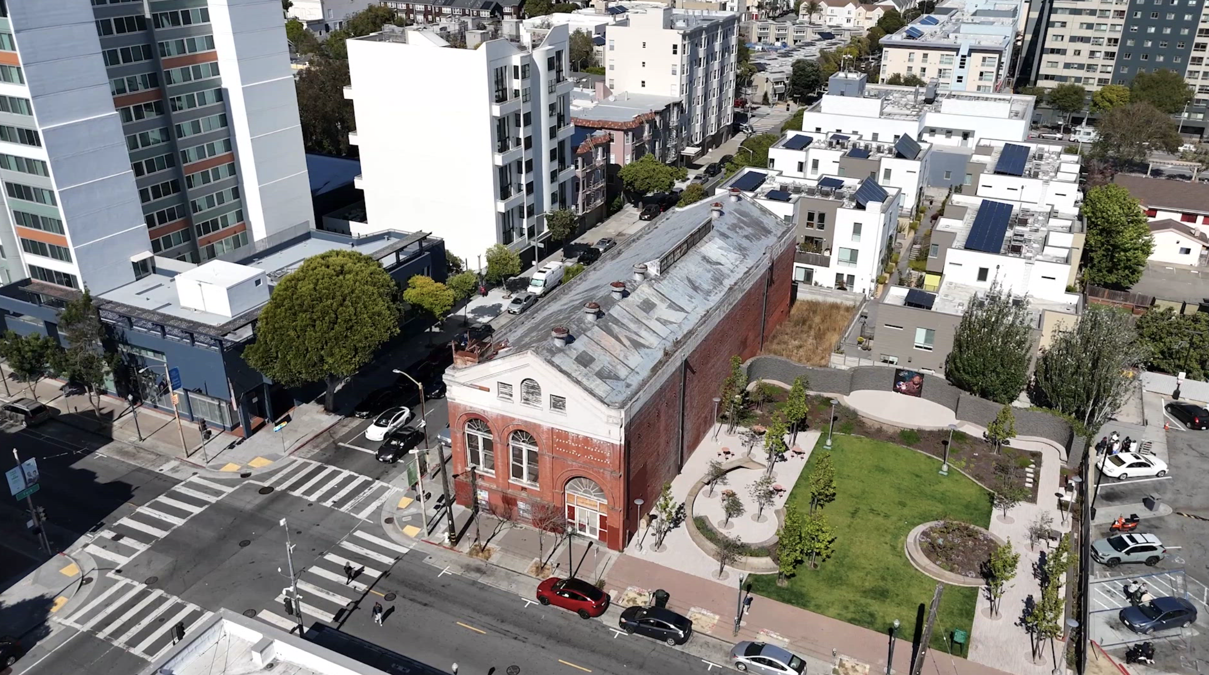 Aerial view of a city block with modern high-rise apartments, a historic red brick building the Fillmore Substation, streets with cars, crosswalks, and a small park with trees and benches.