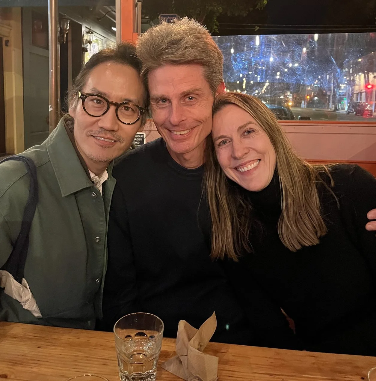 Cadence for Change members, three adults sitting closely together at a table in a restaurant, smiling at the camera with a night city view through the window behind them.