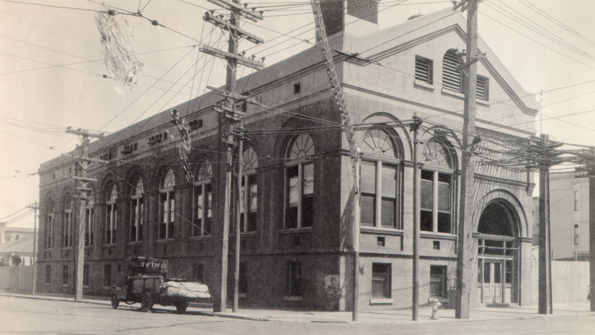 Black and white photo of the Fillmore Substation with arched windows, surrounded by utility poles and numerous overhead electrical wires, with a person riding a bicycle in the foreground.