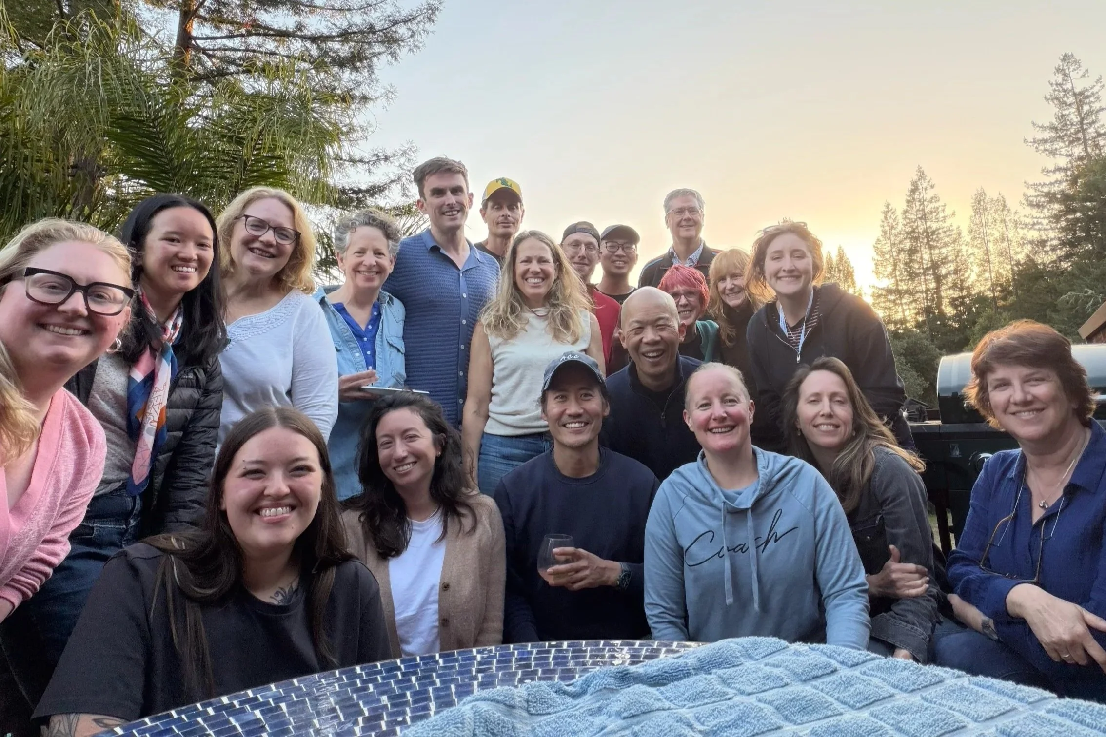 Group of smiling people, Cadence for Change members, gathered outdoors at sunset, with trees and sky in the background.