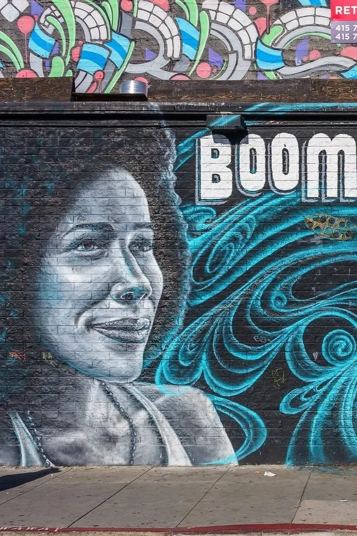 Street art mural of a woman's portrait painted in black, white, and blue on a brick wall, with the word "BOOM" in bold white letters and swirling blue patterns in the background.