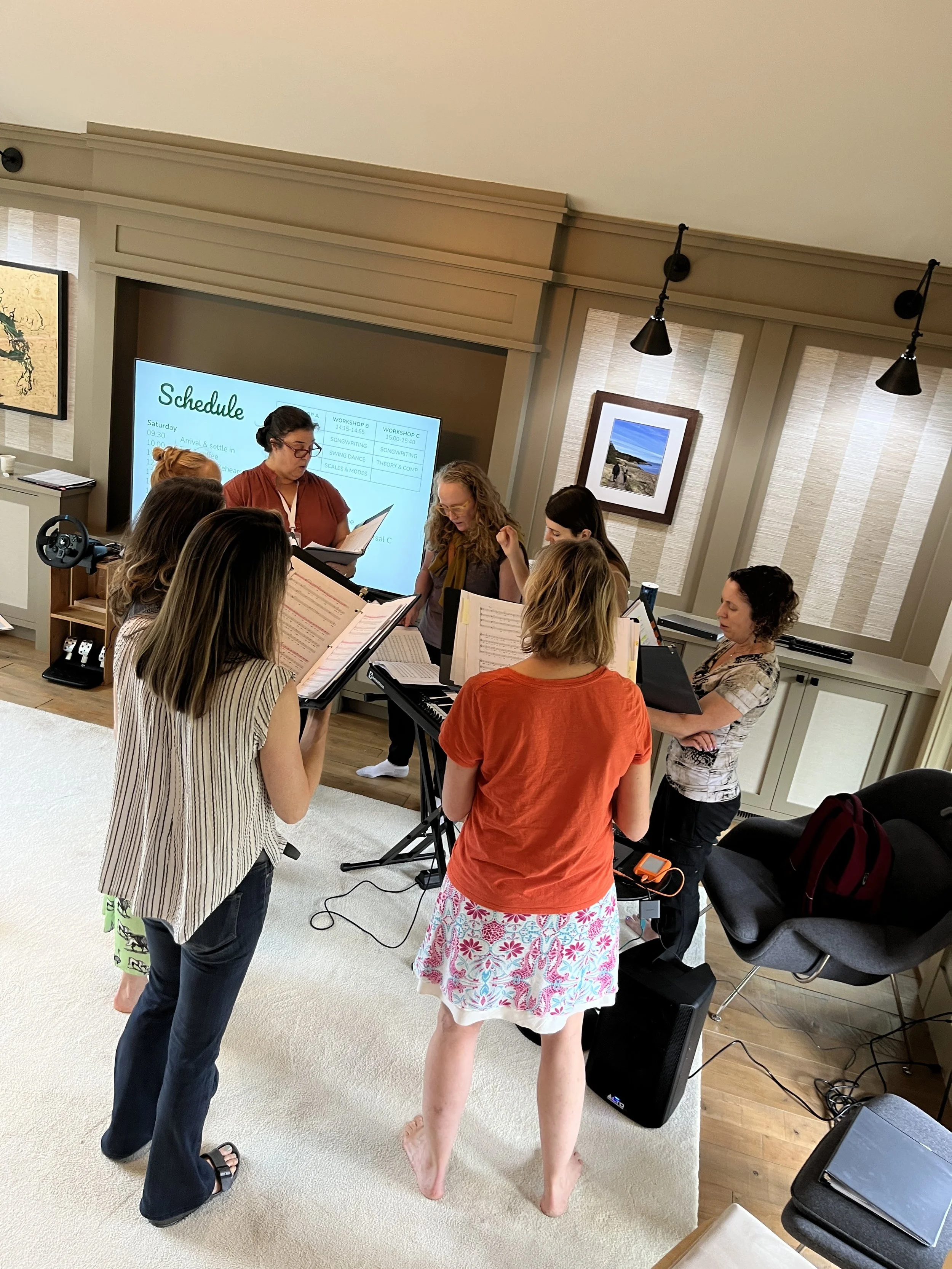  in a room, with some standing around and others sitting at a keyboard. They are engaged in a musical practice or rehearsal. There is a large screen displaying a schedule, framed artwork on the walls, and various electronic equipment and documents around.