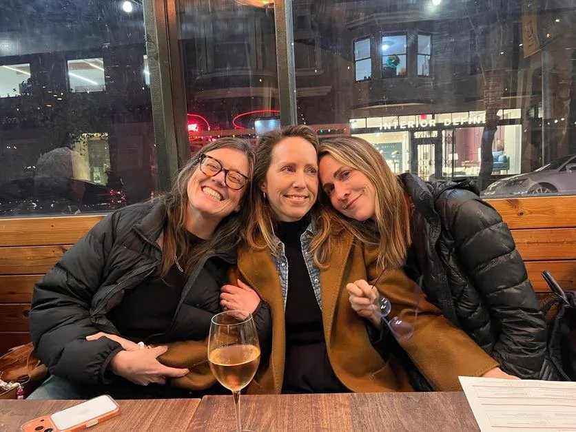 Cadence for Change members sitting together at a restaurant table, smiling and hugging, with a glass of rosé wine in front of them and city lights outside the window behind them.