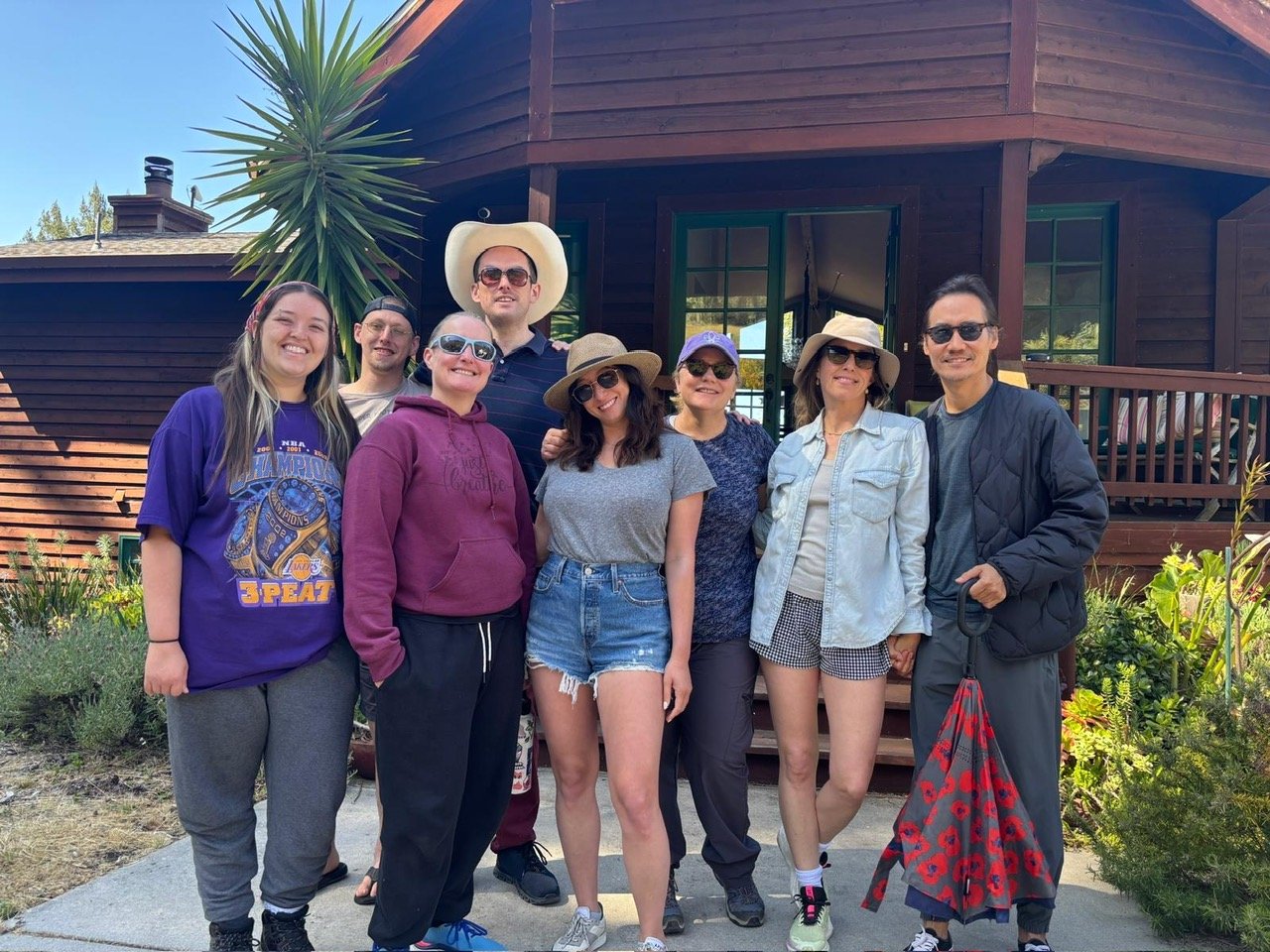 Group of nine people, Cadence for Change members, standing outside in front of a wooden house, smiling, some wearing hats and sunglasses.
