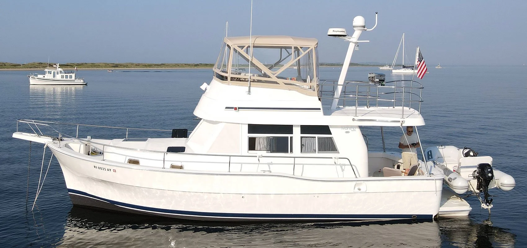 A white motor yacht with a beige covered upper deck, anchored in calm water with a shoreline and other boats in the background; a man is standing near the helm at the rear of the boat.