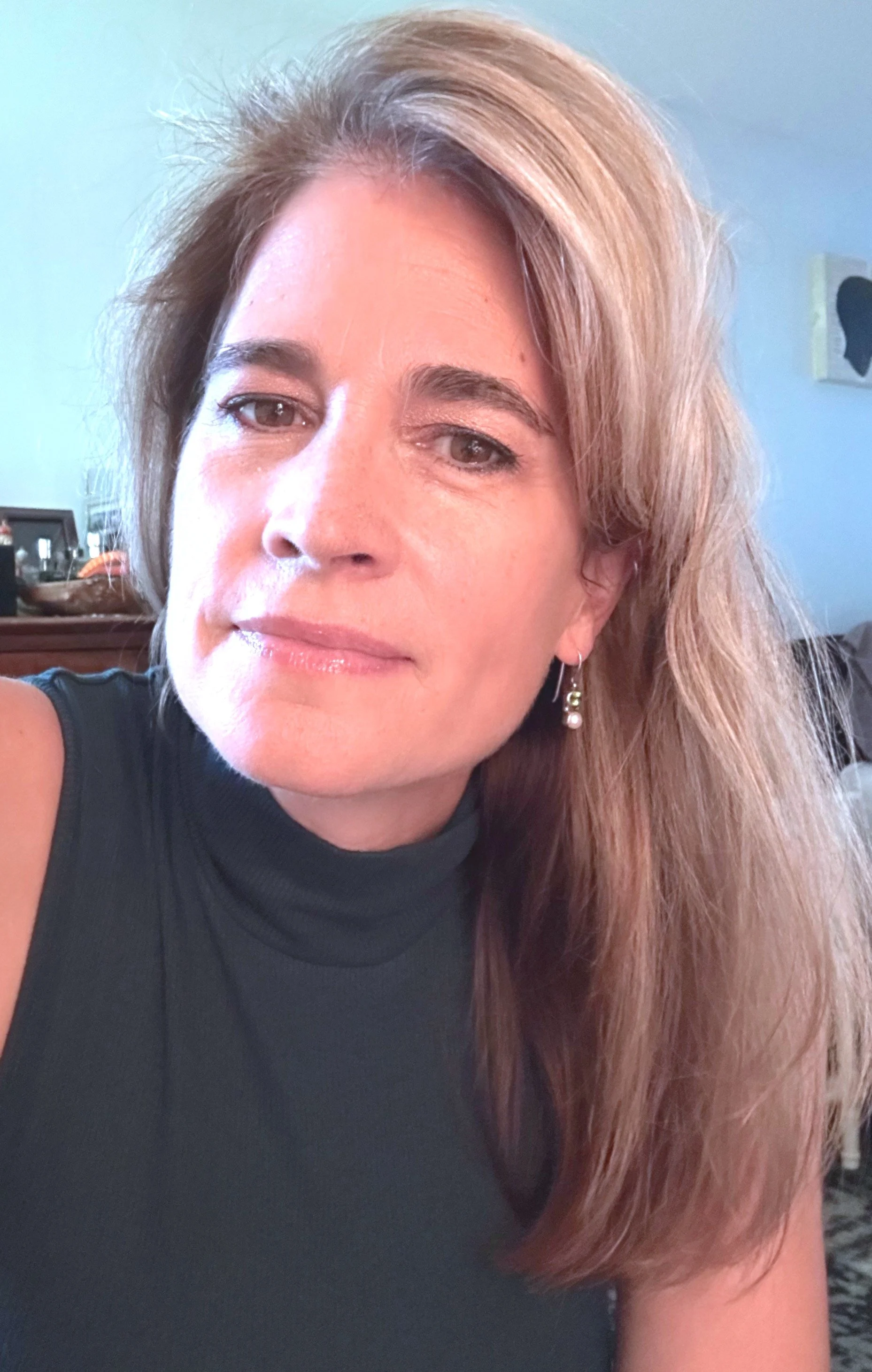 A woman with reddish-brown hair, wearing a black sleeveless top and earrings, posing indoors with a calm expression.