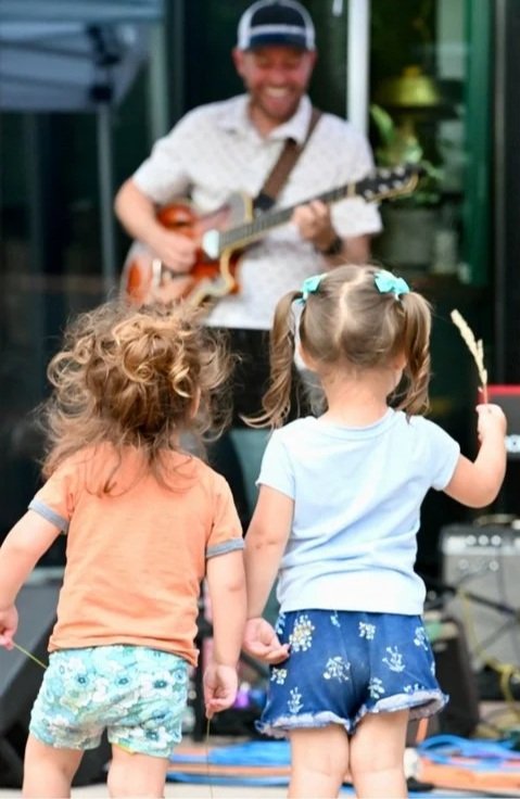 Two young girls with pigtails and colorful outfits watching a street performer playing guitar.