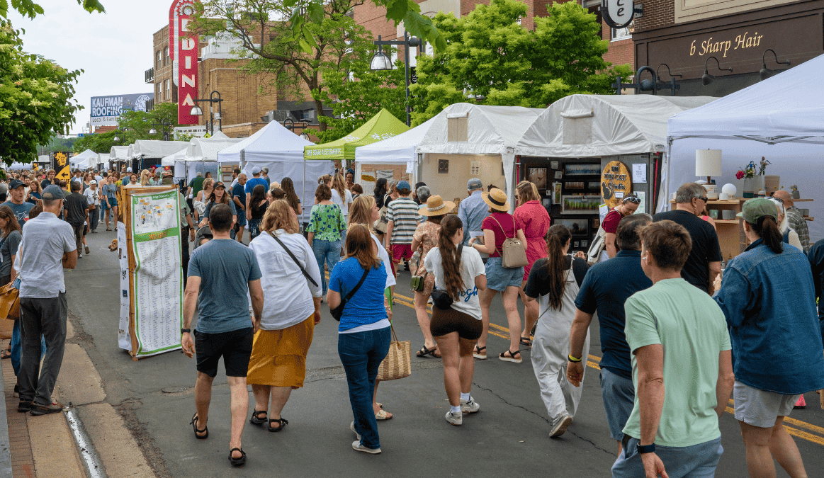 A street fair with white tents and booths, crowded with people walking and browsing, lined with trees and buildings.