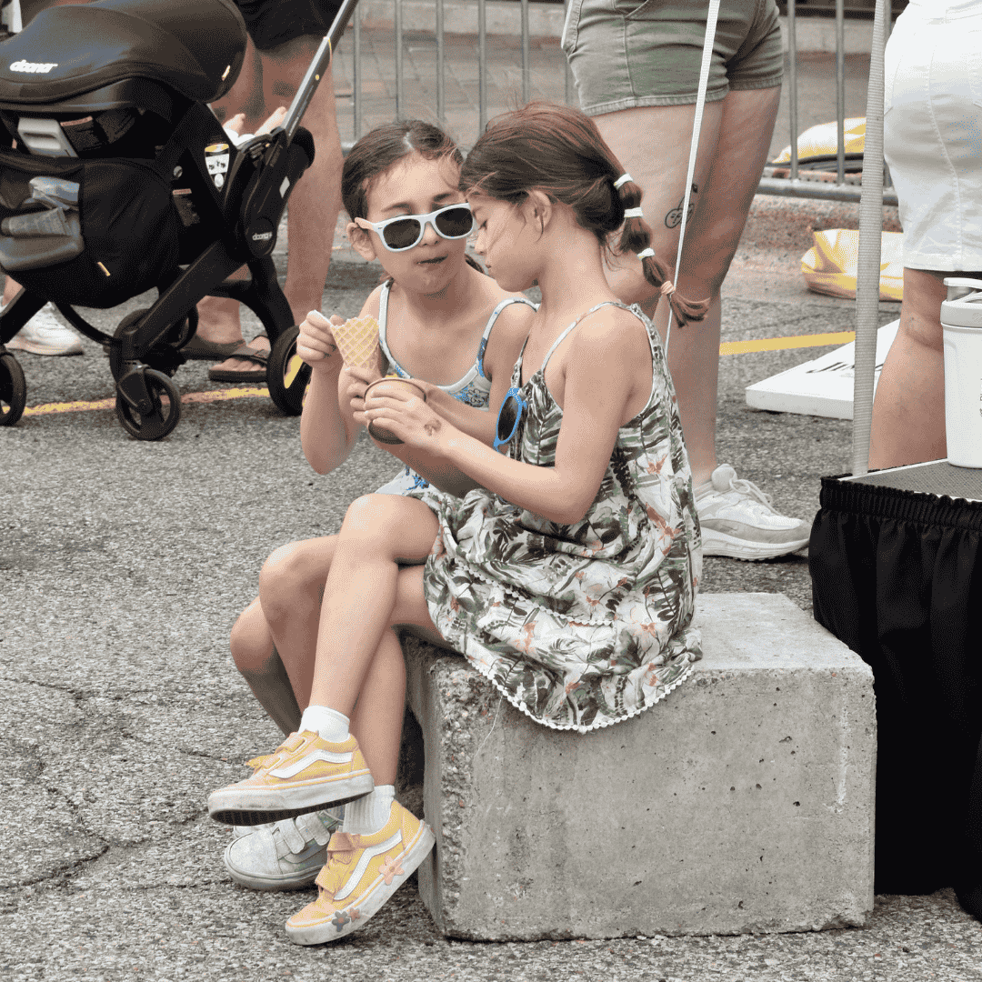 Two young girls sitting on a concrete block, sharing an ice cream, at an outdoor event. They are wearing summer dresses and sunglasses, and are surrounded by other people and strollers.