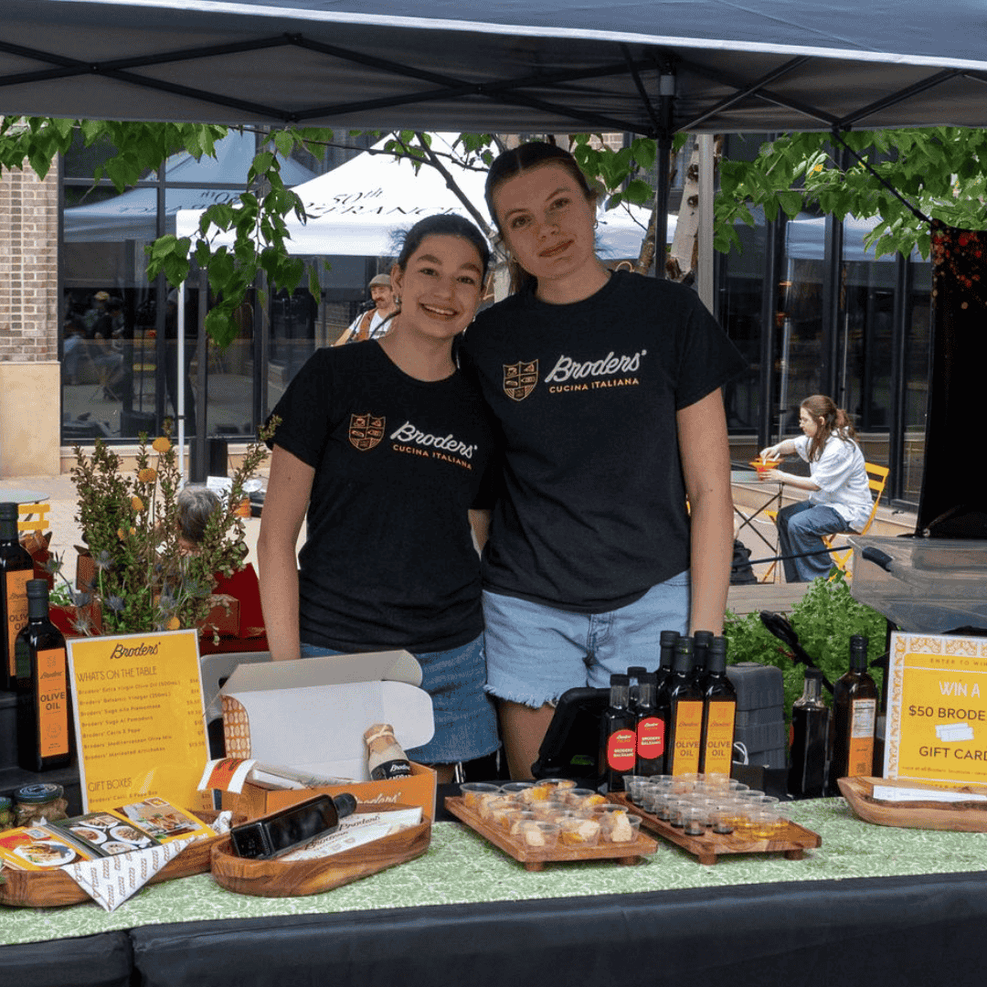 Two women standing behind a table with bottles and packaged products at an outdoor market stall, smiling at the camera, under a black canopy with green foliage overhead.