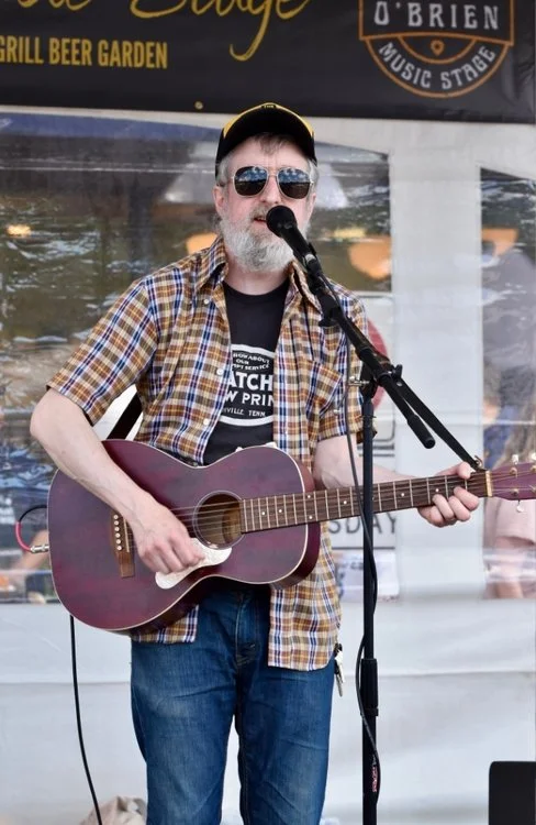 A man with a beard wearing sunglasses, a black cap, and a plaid shirt playing an acoustic guitar and singing into a microphone at an outdoor event.