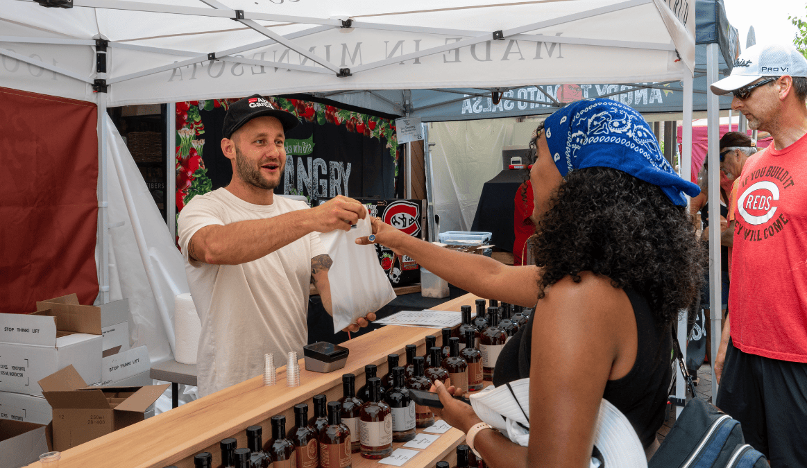 A man in a black cap and white T-shirt handing a paper bag to a curly-haired woman with a blue bandana at a stall selling bottles of sauce or syrup at an outdoor market.