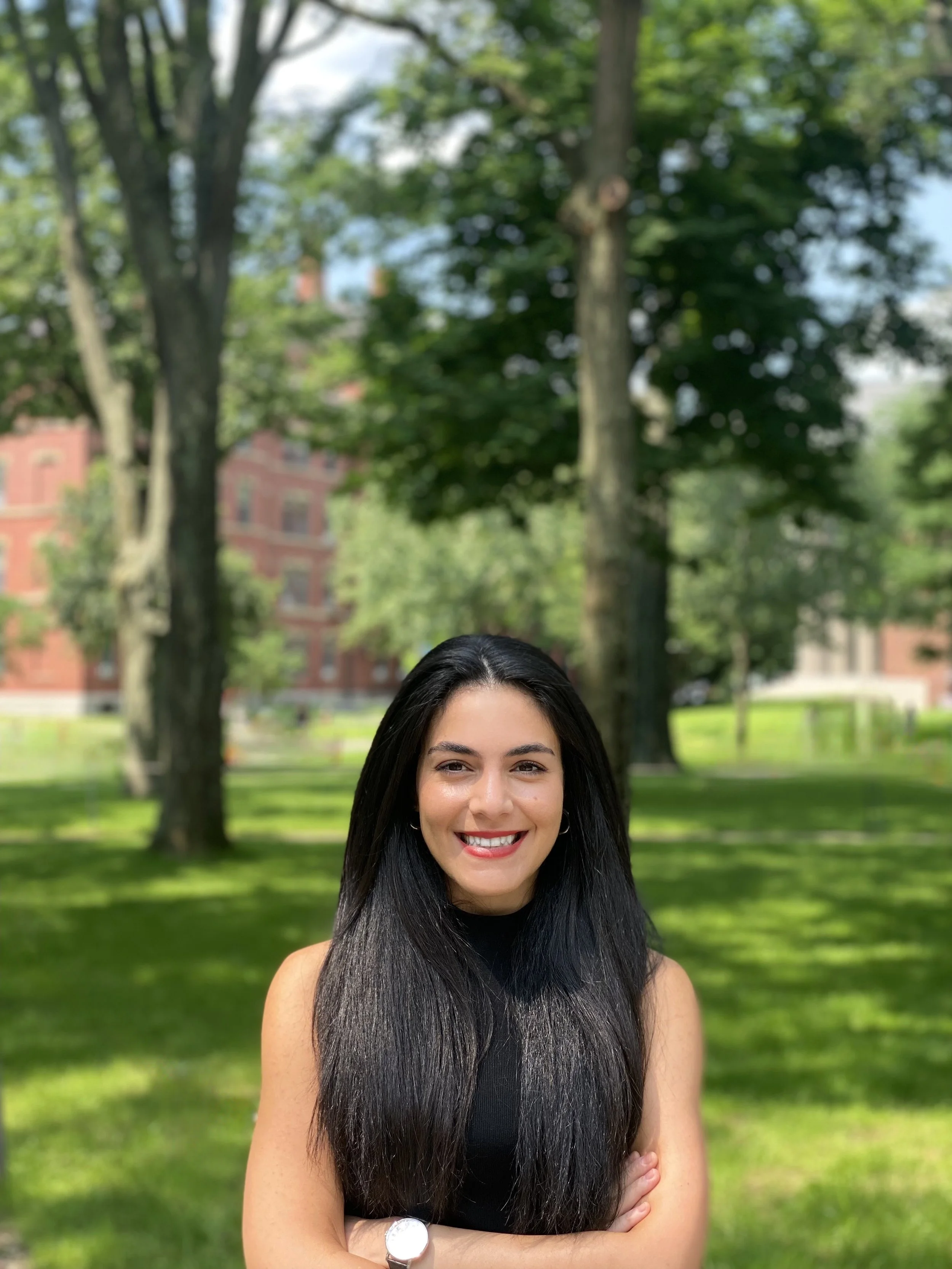 A young woman with long black hair, smiling, standing outdoors in a park with trees and a red brick building in the background.