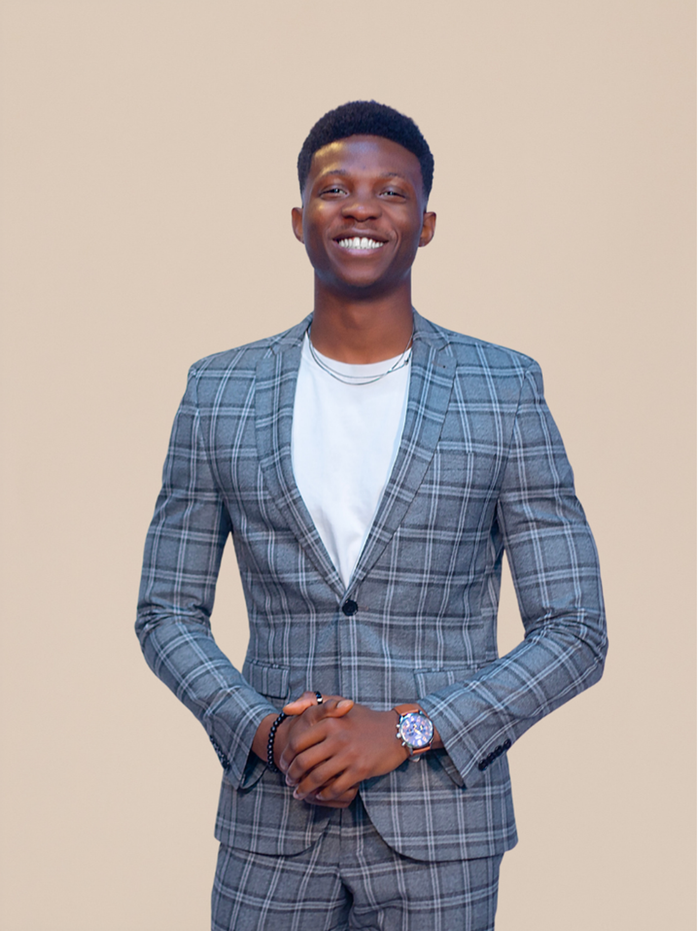 Young man in a gray plaid suit smiling with crossed hands, wearing a white t-shirt, a watch, and a beaded bracelet against a plain beige background.