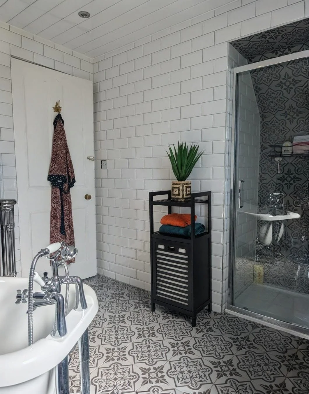 Bathroom with a white brick wall, patterned tile floor, glass shower, and a black plant stand with a green plant and folded towels.