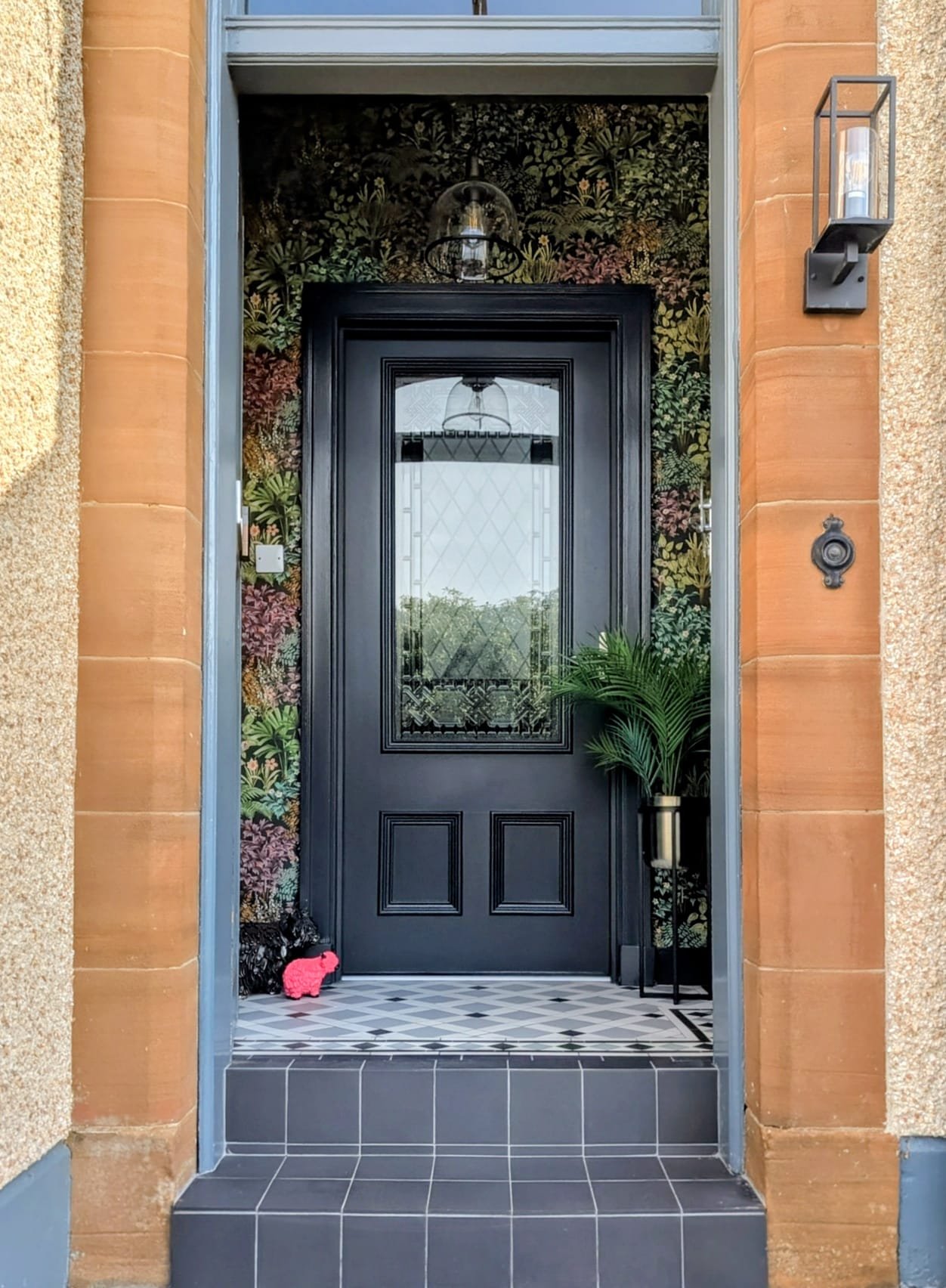 Front entry of a house featuring a grey door with a glass panel, floral wallpaper, a potted plant on a stand, and a pink decorative highland cow figure on the tiled porch.