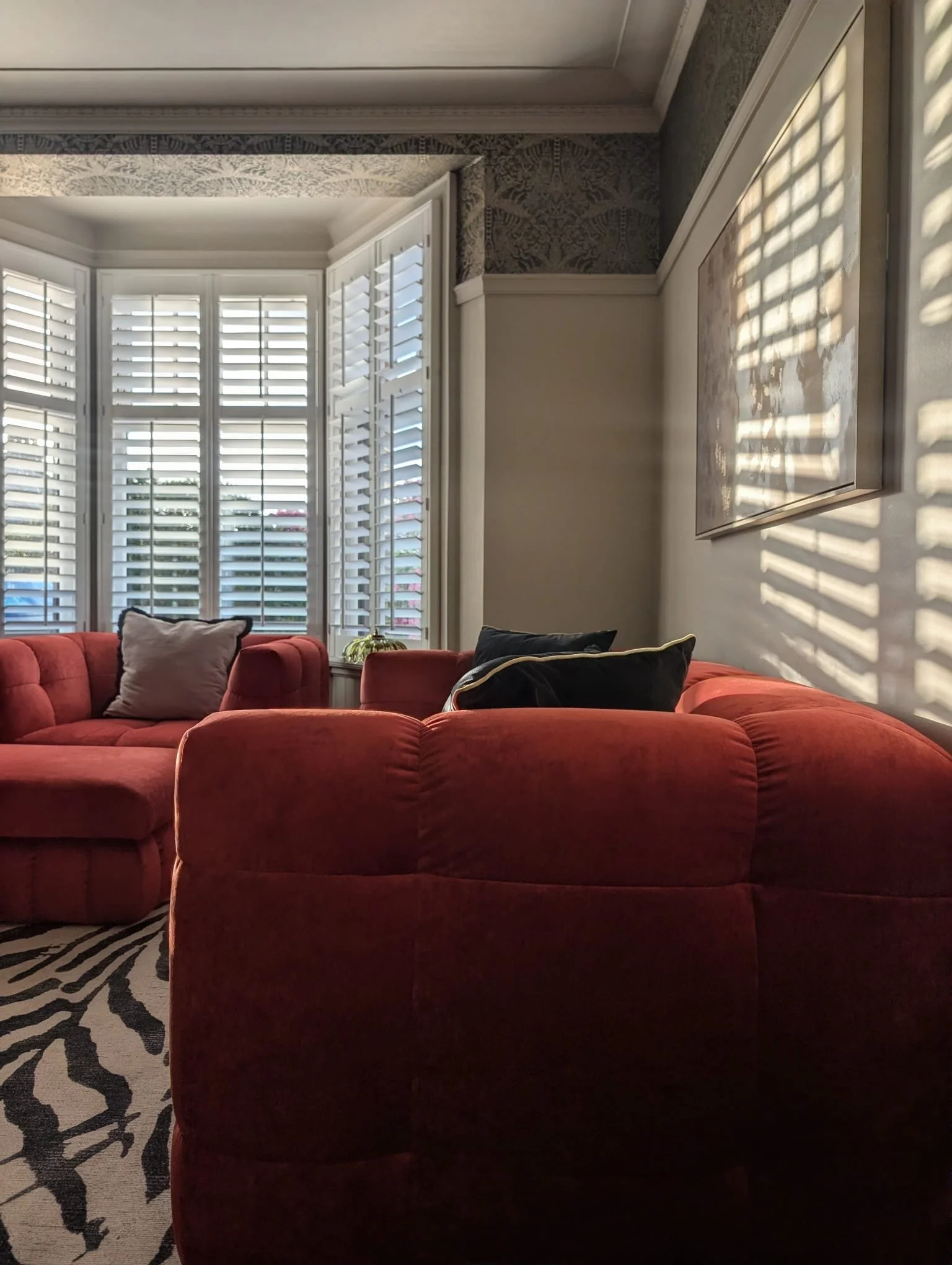 Living room with large windows covered with white plantation shutters, a red sofa with pillows, a patterned area rug, and a wall decorated with framed artwork and patterned wallpaper.
