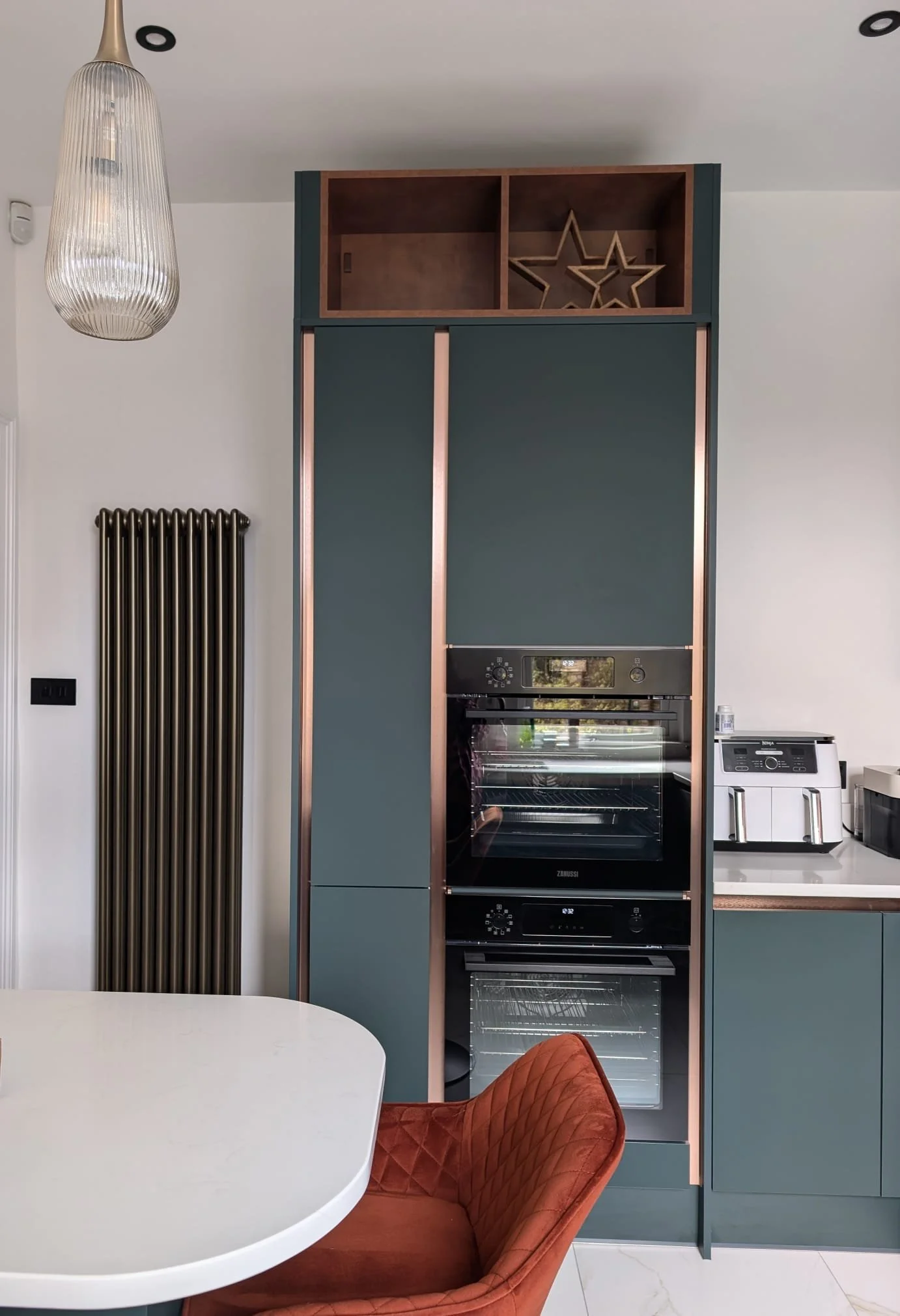 Modern kitchen with built-in oven, green cabinets with rose gold accents, a white rounded table, and a burnt orange quilted chair. Silver star decorative items on open shelves above the cabinets, a hanging glass light, and a black radiator on a white