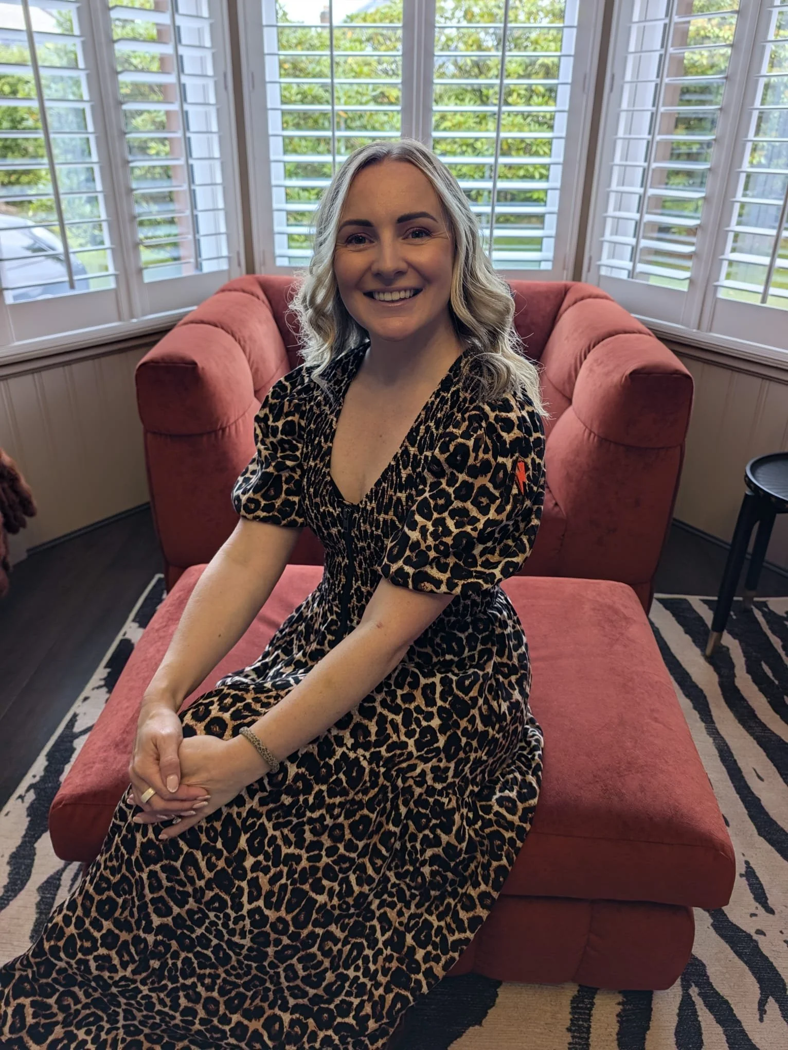 A woman with blonde, wavy hair sitting on a pink couch in front of large windows with white shutters, wearing a leopard print dress, smiling at the camera.