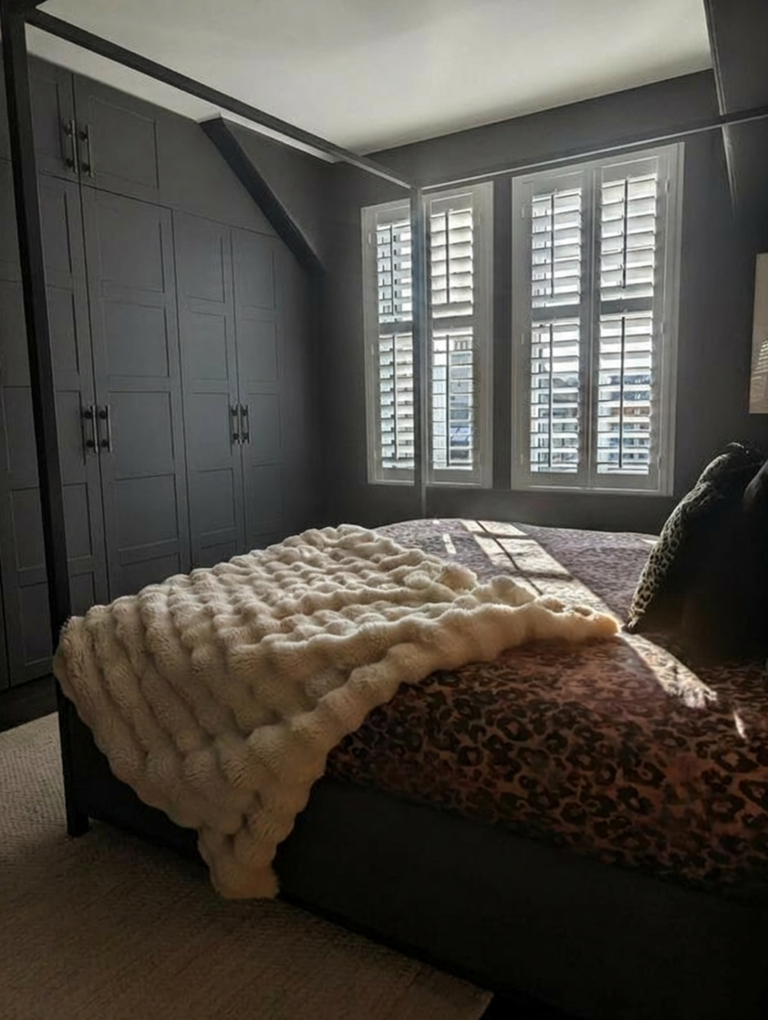 A bedroom with a bed that has a leopard print blanket, a shaggy beige throw, and a patterned pillow. There are three tall windows with white shutters allowing light into the room, and built-in gray cabinets on one side.