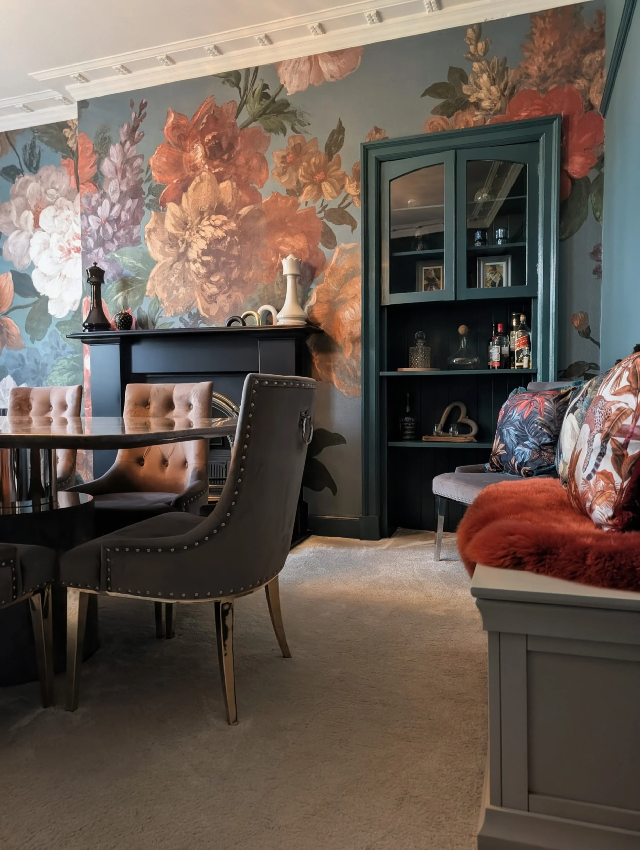 A dining room with a large floral mural on the wall, a dark fireplace mantel with decorative objects, a teal-colored cabinet with glass doors and various bottles inside, a tufted dining chair, and a bench with floral pillow and fuchsia fur throw.