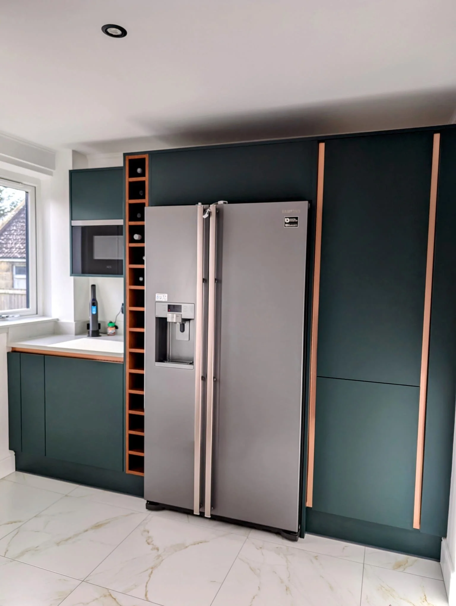 Modern kitchen with green cabinets, a large double-door refrigerator, a window, and a shelf with wine bottle slots. The floor is white marble with gold veining.