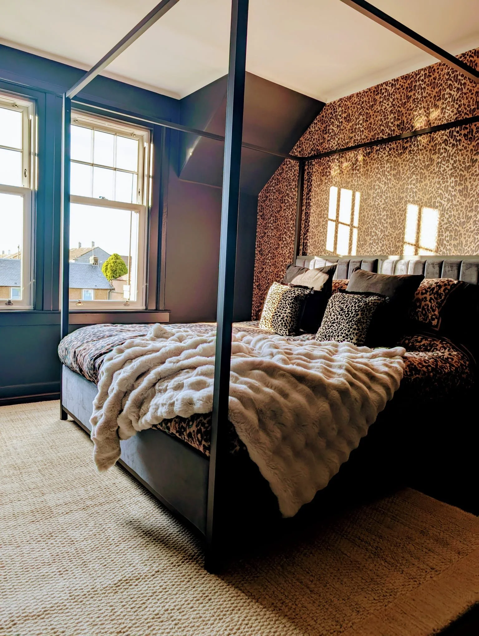A bedroom with a bed covered in a furry blanket, black and animal print pillows, and a leopard print accent wall. The room has a large window with sunlight and a view of neighboring rooftops and a tree.
