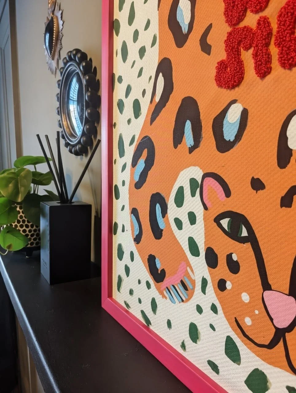 Close-up of a framed modern art piece on a wall, featuring an abstract depiction of a leopard's face with orange, black, green, pink, and white colors, pink framing, and a black background. Part of a black surface with a green potted plant and a blac