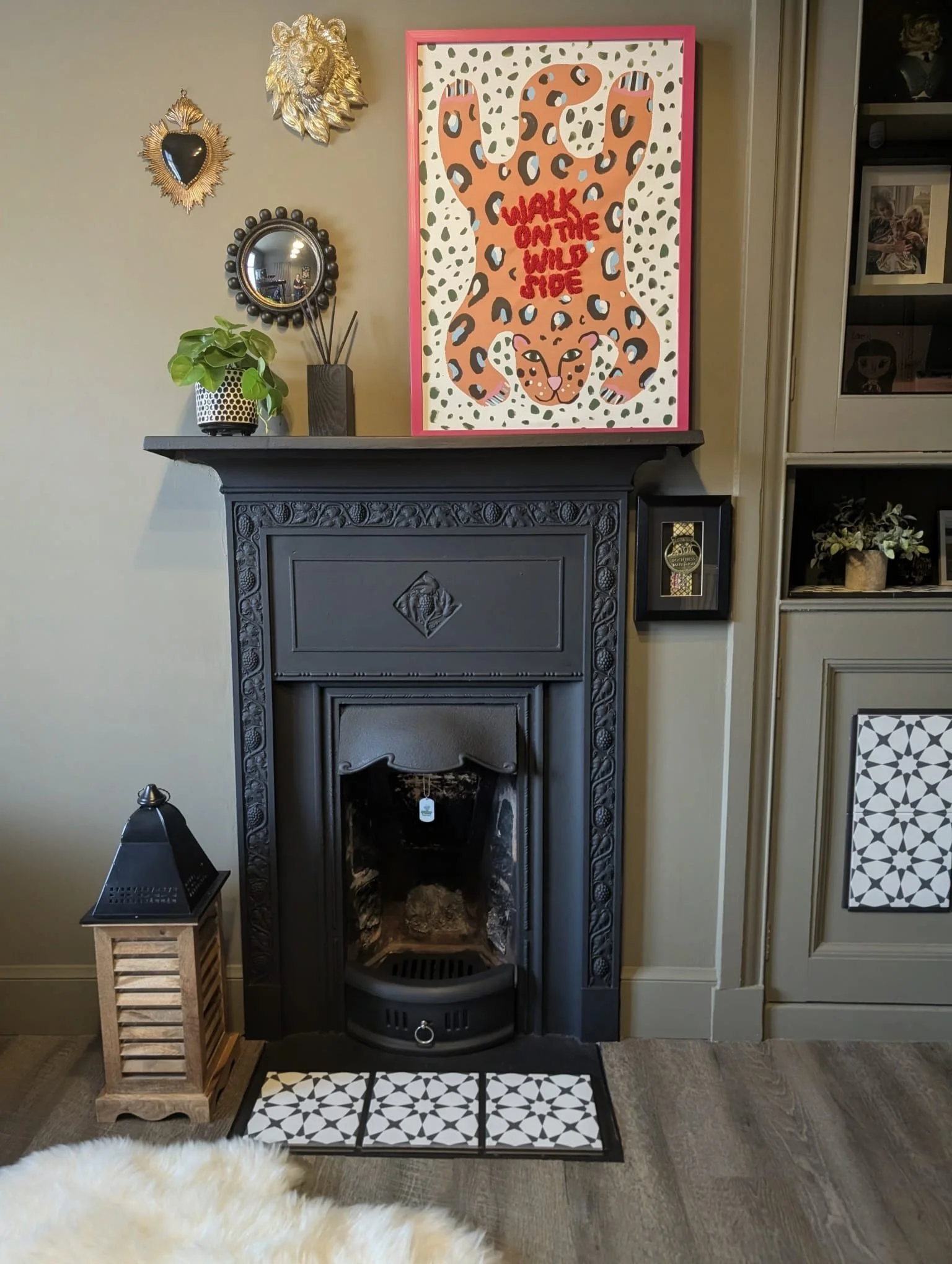A decorative living room corner featuring a dark gray ornate fireplace mantel, with a potted plant and reed diffuser on top. Above the fireplace, there are wall decorations including a golden lion head, a framed mirror, and a black heart-shaped wall 