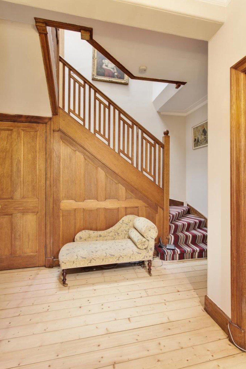 A small vintage-style cream-colored chaise lounge with floral pattern and rounded bolster pillow, placed on a light wooden floor, in front of a wooden-paneled wall and staircase with striped carpeting, in a cozy home interior.